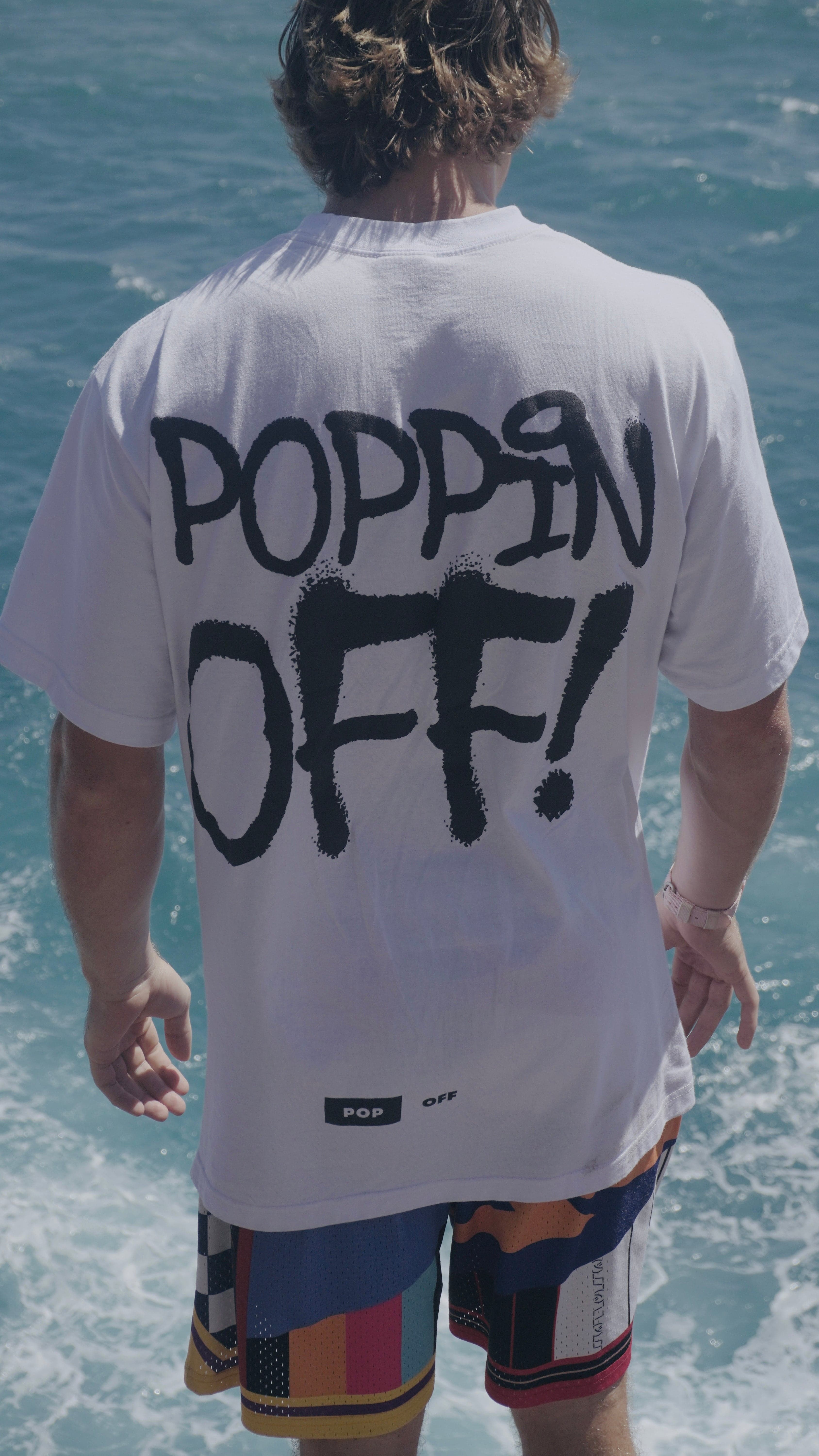 Someone at the shoreline in the white Poppin Off tee, waves behind